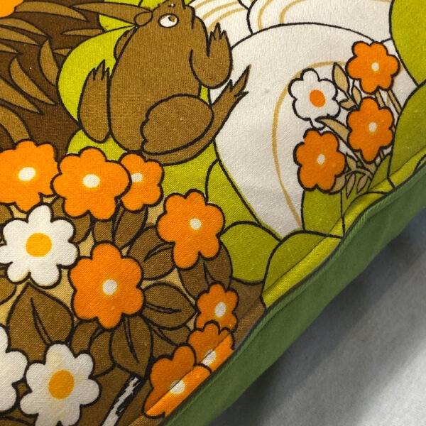 Detailansicht mit Reißverschluss und grüner Hinterseite eines Sofakissens aus 70er Jahre Vintagestoff in hellem Orange, dunklerem Orange, Hellgrün, Karamell, Dunkelbraun und Weiß. Zu sehen ist eine stilisierte Landschaft mit Blumen, Büschen und Bäumen und einem See, bevölkert von Enten, einem Reh, einem Frosch, einem Eichhörnchen, einem Hasen und Vögeln.
