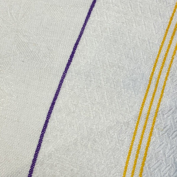 DetailansichtKissen aus strukturiertem weißem Stoff mit zwei lila Längs- und vier Querstreifen, und jeweils 3 gelben Streifen an der Seite, gefolgt von mehreren lila Streifen