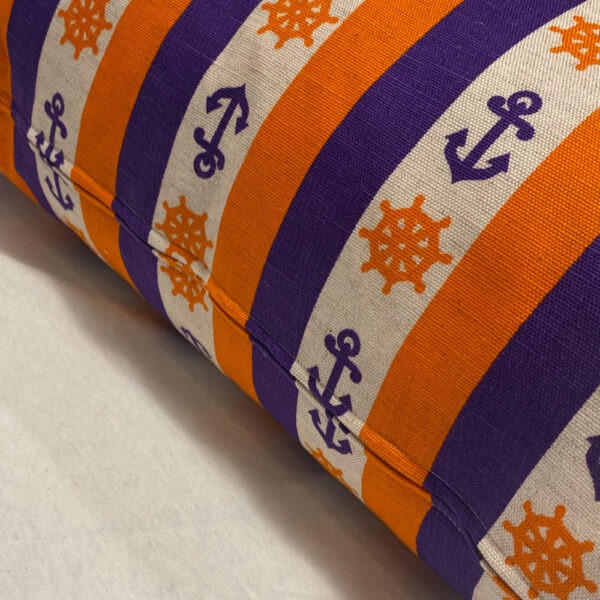 Detailansicht mit Reißverschluss eines Sofakissens aus einem lila, orange, cremeweiß gestreiften Stoff. Die Streifen sind regelmäßig, wobei im cremeweißen Streifen lila Anker abwechselnd mit orangen Steuerrädern eingewebt sind